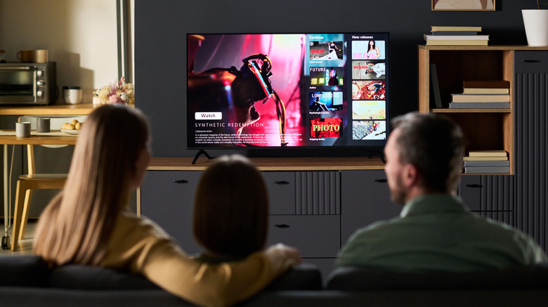 An adult man and woman have a small child sitting between them as they're seated on a couch watching TV, the television is displaying what appears to be a fake home screen for a streaming service