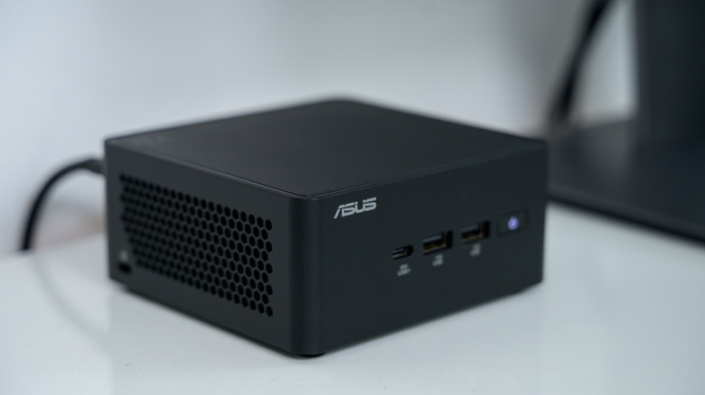 A black ASUS mini PC sitting on a white surface, it has one cable connected to it that's viewable in the photo, and its blue power light on the front is glowing