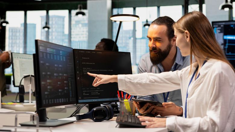A woman with two monitors in front of her pointing at the secondary monitor, showing something to her colleague