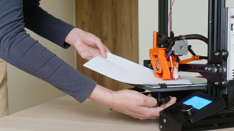 A person checking the 3D printer's bed leveling with the help of a sheet of paper