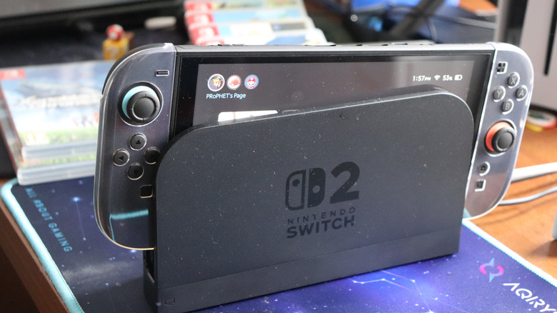 A Nintendo Switch 2 partially out of it dock as it sits on a table, several game boxes are behind the system