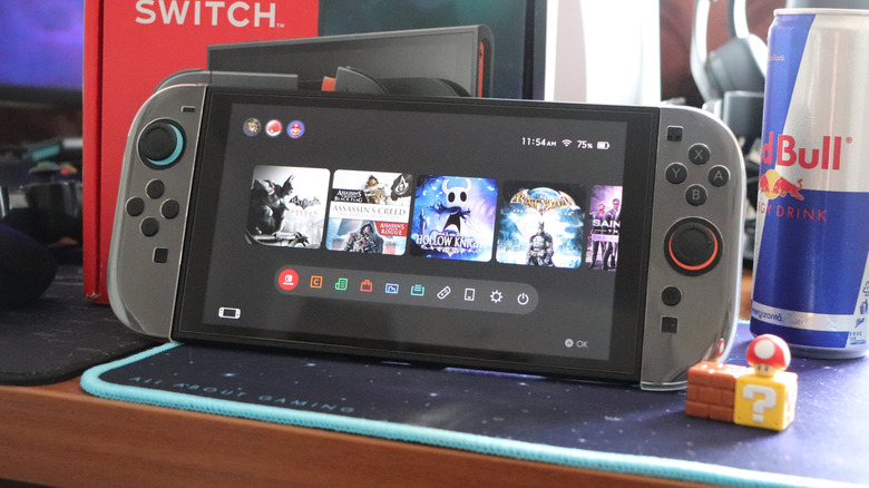 A closeup of the front of a Nintendo Switch 2 displaying its Home screen, the console sits next to a can of Red Bull, and the Switch's packaging sits behind the system
