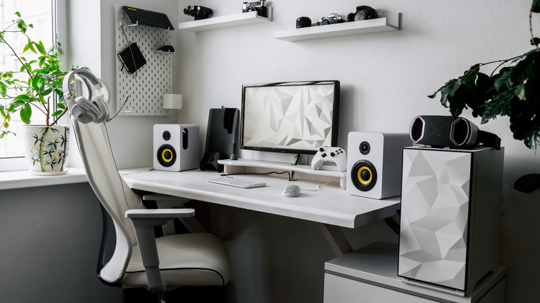 Two white speaker enclosures with black speakers sit between a monitor, gaming controllers, laptop, keyboard, and mouse, multiple accessories surround the rig as the items hang from the wall or sit on white shelves