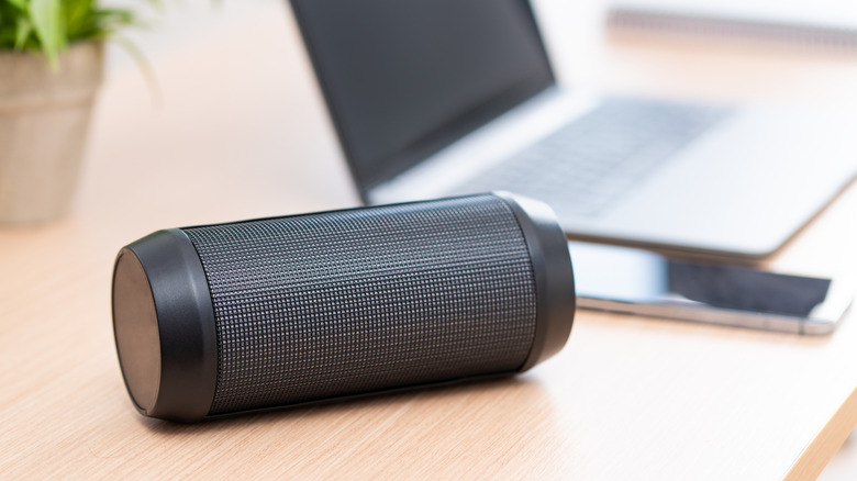 A black wireless speaker sits next to a smartphone and a laptop, all three items sit atop a wooden desk with a potted plant