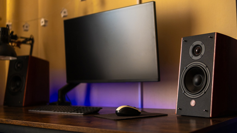 A pair of black speakers with wooden trim sit between a black monitor, a keyboard, a gaming mouse, and a mousepad, there is a faint hint of blue seen coming from behind the desk
