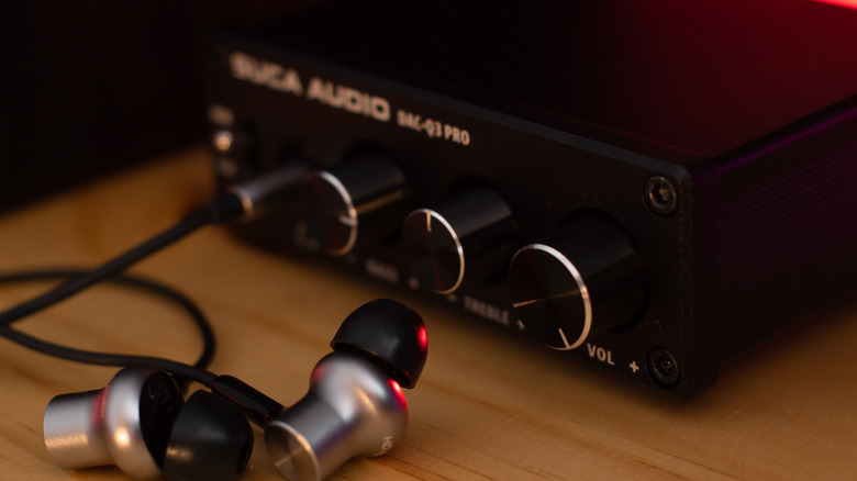 A pair of silver and black wired earbuds sit next to a digital-to-analogue converter (DAC), both objects are sitting on a wooden surface