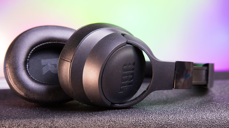 A pair of black JBL headphones (seemingly wireless) sit on a black vinyl surface, a blend of green and purple hues serves as the backdrop