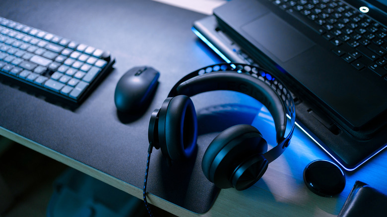 A pair of black wired headphones sit next to a black laptop, black mouse, and black keyboard