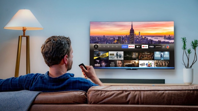An individual sitting down on a couch holding an Amazon Fire TV remote in their right hand, they are facing an Amazon Fire TV that is displaying its Home screen