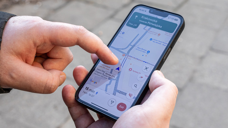 An individual holding a smartphone in their right hand points to the navigation icon on the Google Maps screen with their left hand