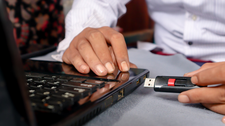 A man's hand holds a USB flash drive connected to a laptop on a table.