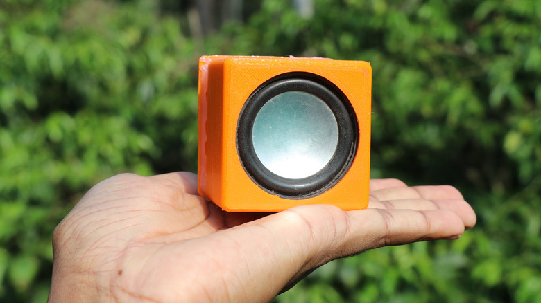 A closeup of an individual's left hand as they hold a small orange speaker, the photo was taken outdoors, as several bushes cover the background