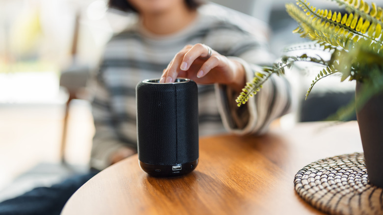 Two fingers from an individual in a striped shirt's left hand touch the top of a black Bluetooth speaker, the speaker itself sits on a brown table next to a potted plant