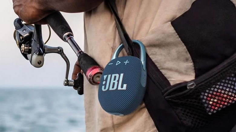A silber and black JBL Clip 5 is attached to a black bag being carried by an individual holding a fishing pole, the photo focuses on the speaker, leaving much of the individual obscured
