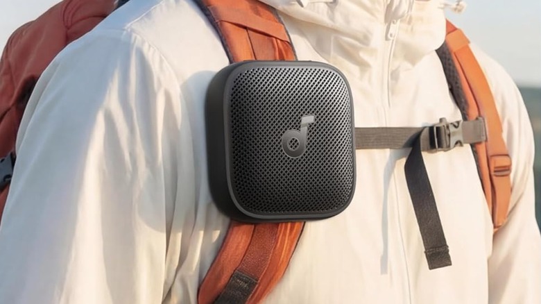 A black Soundcore Boom Go 3i is attached to a user's orange backpack, the user is wearing a white jacket and their head is unable to be seen