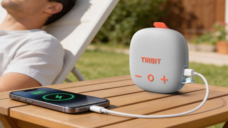 A grey Tribit StomBox Micro 3 is connected to a smartphone by a USB-C cable, both objects sit on an outdoor table next to a person in a white shirt