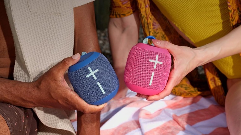 A pink and a blue Ultimate Ears Wonderboom 4 are being held next to each other by different different individuals, the photo is a closeup of the speakers so the faces of the individuals cannot be seen