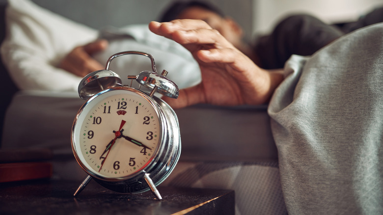 A person in bed reaches over to an analog alarm clock on a bedside table.