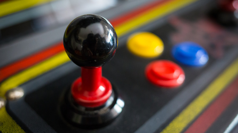 A closeup of an black and red arcade stick next to yellow, blue, and red arcade buttons, the items appear attached to an arcade cabinet