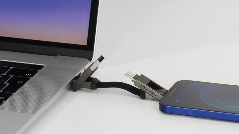The Rolling Square inCharge Universal Cable Charger Keychain, the item is attached to a MacBook and an iPhone, with the charging cable sitting between these two Apple items