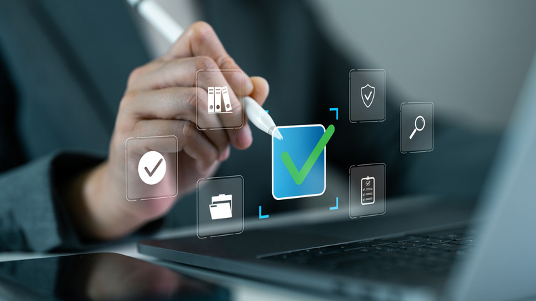 Hand holding a stylus over a laptop with digital icons all around.