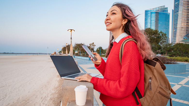A woman using a smartphone and laptop by a beach.