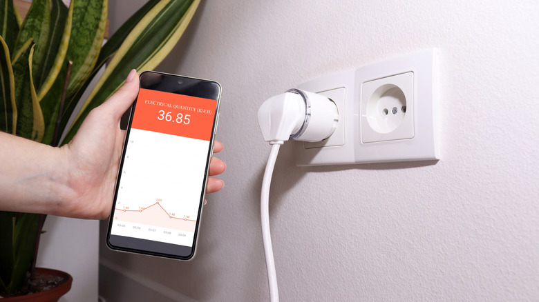 Hand holding smartphone displaying energy monitoring app next to smart plug in wall.