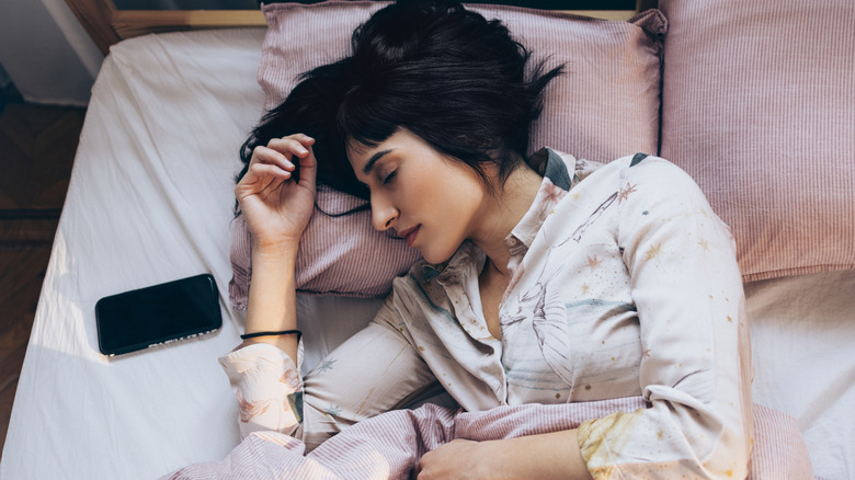 An individual lying in a bed with eyes closed and a smartphone next to them