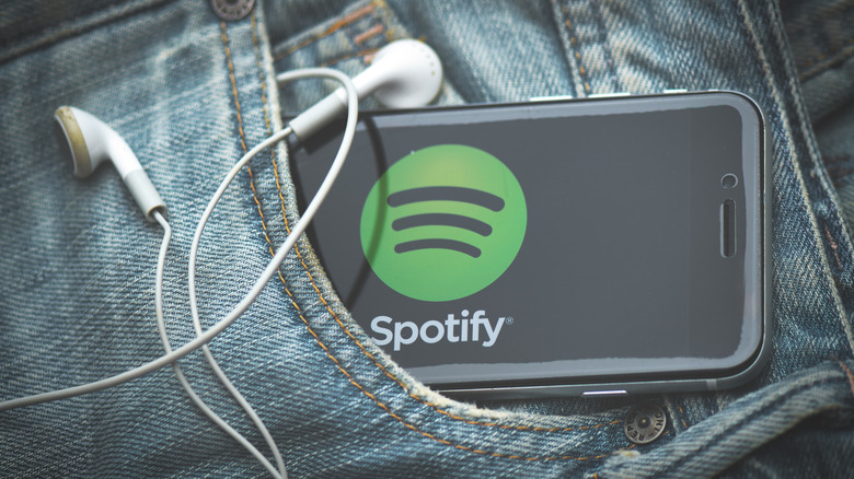 Corded earbuds and a smartphone displaying the Spotify logo hang out of a jeans pocket