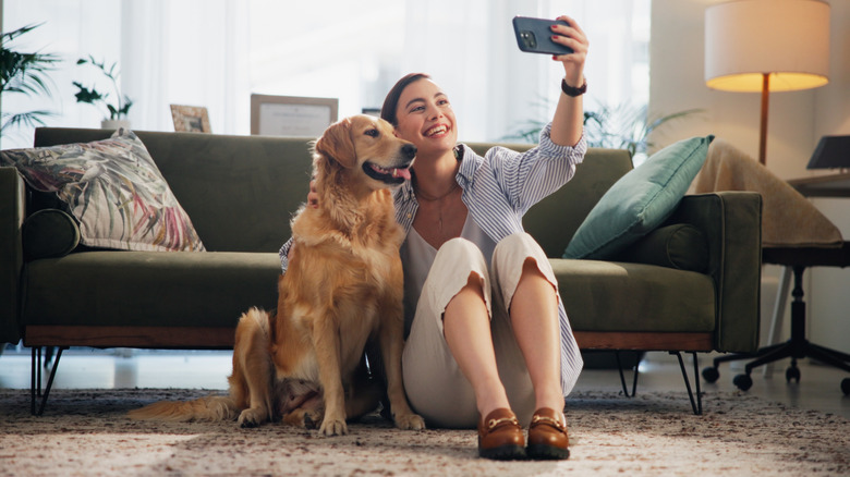 An individual sits on the ground in front of a green couch as they take a selfie on a cellphone with a smiling golden retriever