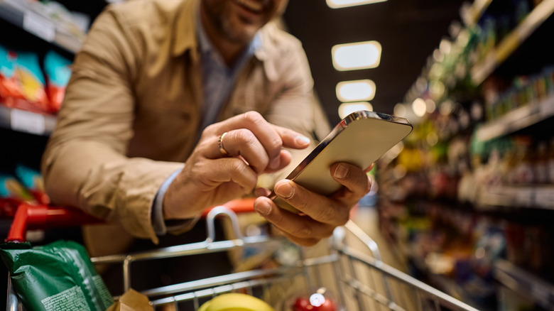 An individual wearing a gold ring on their right hand is checking something on their smartphone as they lean over a shopping cart filled with groceries while in a store