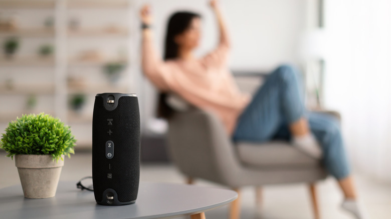 A Bluetooth speaker on a desk in the foreground and a woman dancing while sitting on a chair in the background.