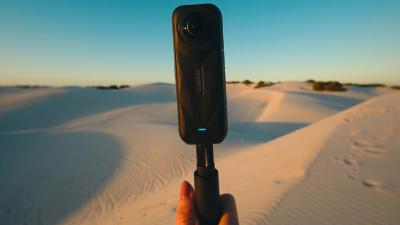 An Insta360 X5 360-degree camera is held up in front of a sprawling landscape of sand dunes.