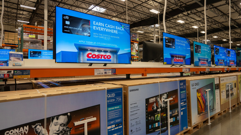 Televisions displayed in a row inside a Costco warehouse, with large TV boxes stacked below and promotional screens above