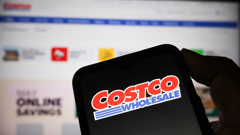 A smartphone displaying the Costco Wholesale logo in front of a screen displaying the Costco website