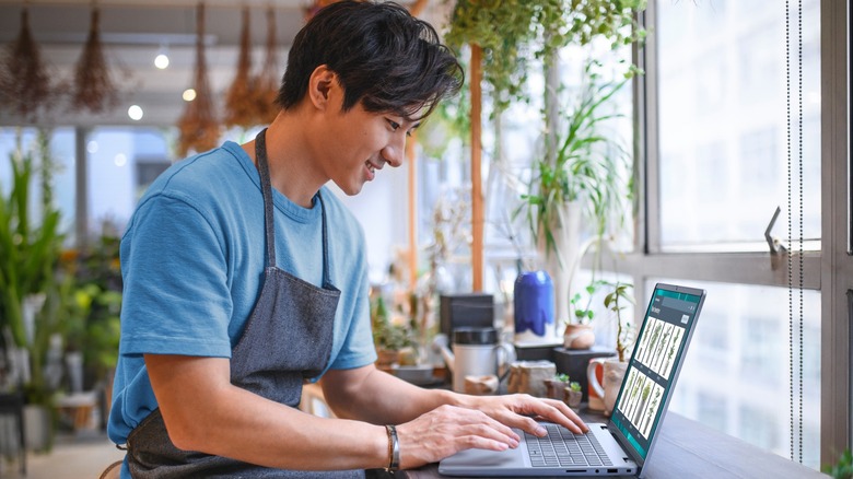 An individual in a blue shirt and darker blue smock sits at Dell 16-inch Touchscreen Intel Core Laptop, the person appears to be in a plant store