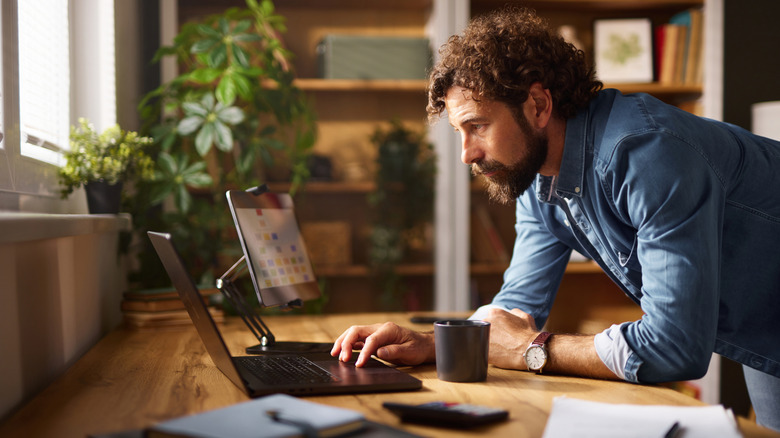 An individual in a blue longsleeve shirt stands over a desk as they have their right hand on a black laptop, there is also a black tablet on a stand next to the person