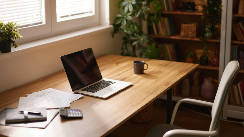 A silver laptop, likely something from the MacBook lineup, sits at a desk next to a black coffee cup, a calculator, a journal, and some papers, no one is sitting at the desk