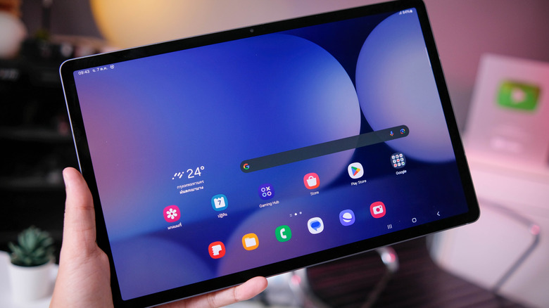 A Samsung Galaxy Tab S10+ being held in a user's left hand against a blurred background, a chair, a plant, and what appears to be a YouTube trophy can be seen in the background
