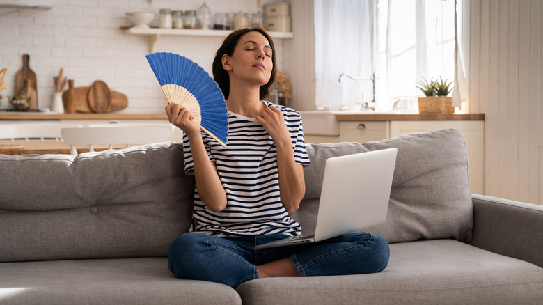 A woman fanning herself while sitting on a living room sofa with a laptop on her lap