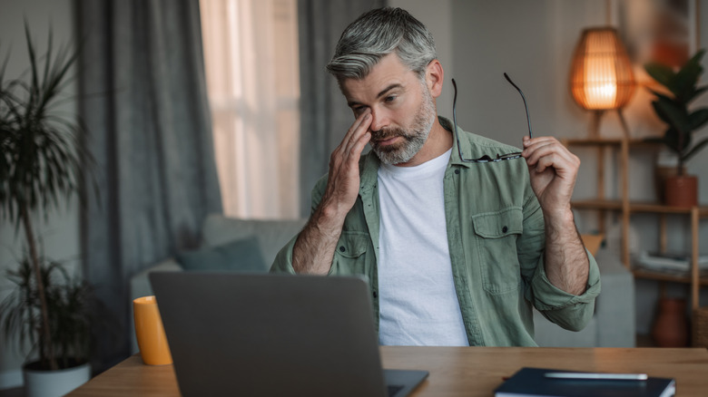 A man with grey hair and a beard takes off his glasses to look at his laptop screen with a stressed expression on his face