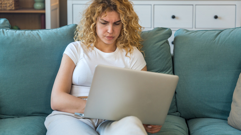 A woman tries to use a laptop on her lap while sitting on a sofa.