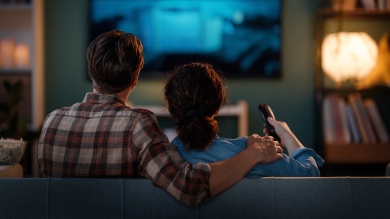 An individual with their right arm around someone else's shoulder as they sit and watch TV, the image is taken from behind and the second individual has a remote control in their right hand