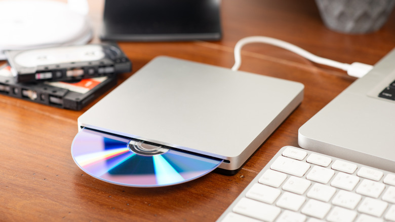 A blue compact disc sits half way protruded from a disc reader that's connected to a silver laptop, a white keyboard and two cassette tapes can also be seen in the image