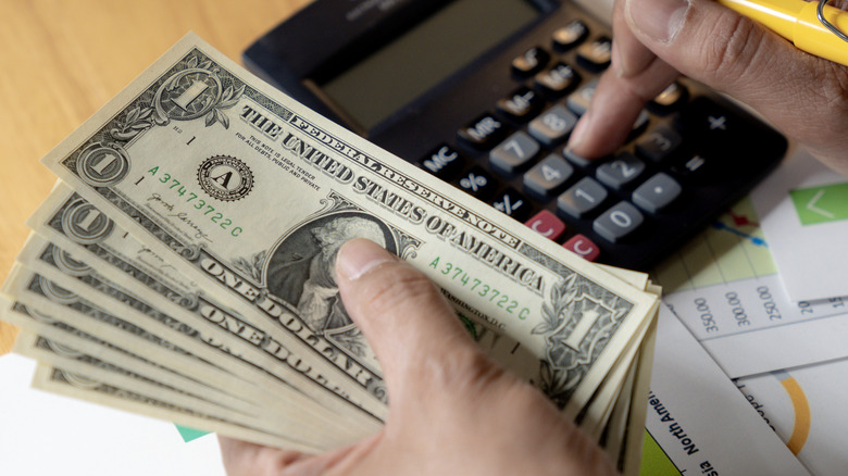 A person holding U.S. currency and typing numbers on a calculator