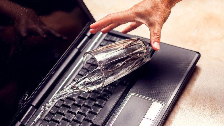 A hand dropping a glass of water on a laptop with water being spilled all over the keyboard