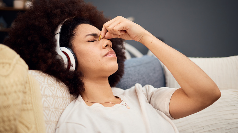 An individual in a white shirt sits on a white couch and pinches the top of their nose with their left hand, the person is wearing silver headphones