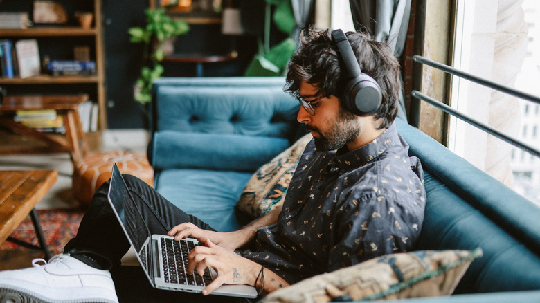 A person in a dark blue shirt sits on a couch inside what appears to be a trendy coffee shop or a hip apartment, the person is typing on a laptop as they wear black headphones