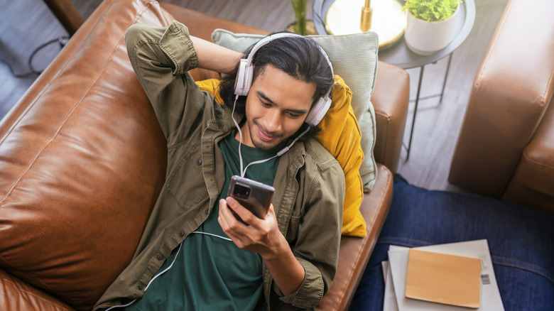 An individual in a light green shirt with a darker green shirt underneath sits on a brown leather couch as they stare at a smartphone held in their right hand, the person is wearing white headphones