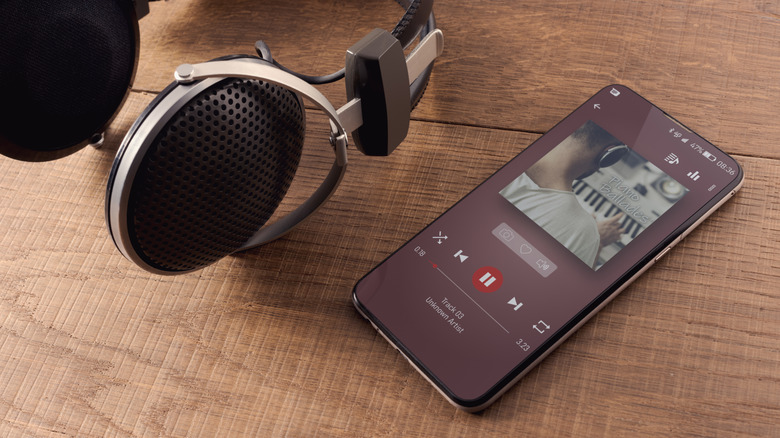A smartphone displaying a generic music player sits on a hard surface next to a pair of black and silver headphones